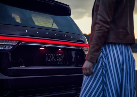 A person is shown near the rear of a 2024 Lincoln Aviator® SUV as the Lincoln Embrace illuminates the rear lights | Magic City Lincoln in Roanoke VA